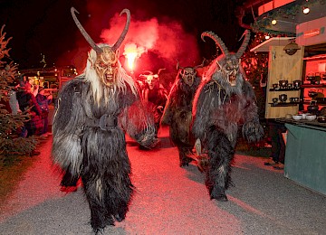 A frightening Krampus mask with wild teeth, red and black tones, and shaggy fur dominates the image. Twinkling lights in the background suggest a festive, pre-Christmas event. The scene captures the traditional Alpine Krampus figure, known as the dark companion of St. Nicholas, bringing excitement and a touch of fear to holiday markets.