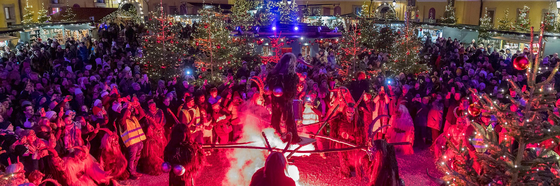 The image captures a festive musical performance at the Hellbrunn Advent Market in Salzburg. In the foreground, four musicians are seated on a cozy, Christmas-themed stage, framed by decorated Christmas trees adorned with red ornaments and warm lights. They are playing traditional holiday music for a cheerful crowd gathered in the courtyard. In the background, visitors stroll between illuminated market stalls, some with strollers and children, all enjoying the lively ambiance. The scene radiates warmth, community, and the joy of music, perfectly complementing the traditional charm of this picturesque Christmas market setting.