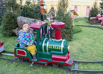 A child joyfully rides a small, colorful train named "El Paso" through a snowy winter landscape. Surrounded by decorated Christmas trees and festive decorations, the scene radiates holiday spirit. It captures the magic of Christmas, childlike excitement, and cozy family moments at a charming Advent market.